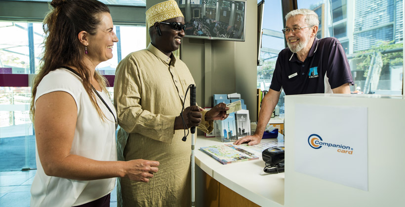 A man and his companion stand at a counter with a man on the other side. The pair are using a Companion Card.