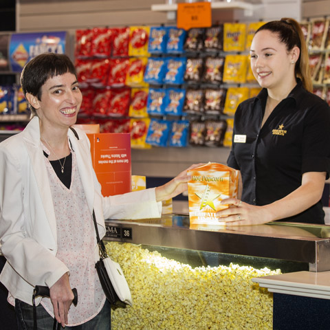 A woman stands at the candy bar of a cinema, while the cinema employee hands her a box of popcorn.